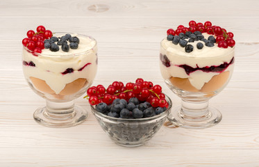 Sweet dessert in glass with biscuits, cream, currants and blueberries on wooden background.