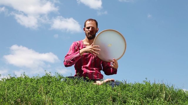 Young man playing the tambourine sitting on the green grass on blue sky background