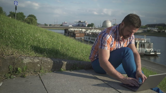 Man writing on a laptop on the stairs by the river, pan shot, right
