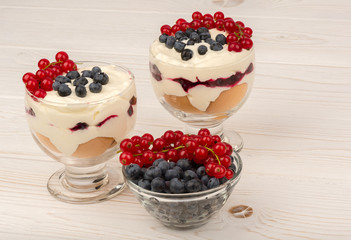 Sweet dessert in glass with biscuits, cream, currants and blueberries on wooden background.