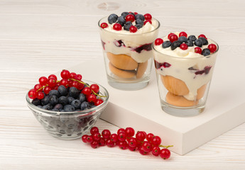 Sweet dessert in glass with biscuits, cream, currants and blueberries on wooden background.