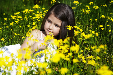 Little girl with dark hair sitting on a field of of yellow flowers on the background of green grass