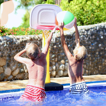 Two happy teenage boys, sportive twin brothers, having fun together playing basketball with colorful inflatable ball in outdoors swimming pool in aquapark during summer sea vacation in tropical resort