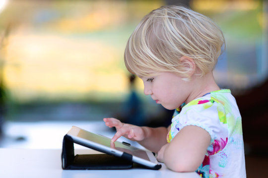 Happy little child, adorable blonde toddler girl enjoying modern generation technologies playing indoors using tablet pc with touchscreen