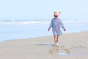 Cute active child wearing pink sunglasses playing and running on wide sandy beach. Happy little girl enjoying summer holidays on a sunny day. Family with young kids on vacation at the North Sea coast.