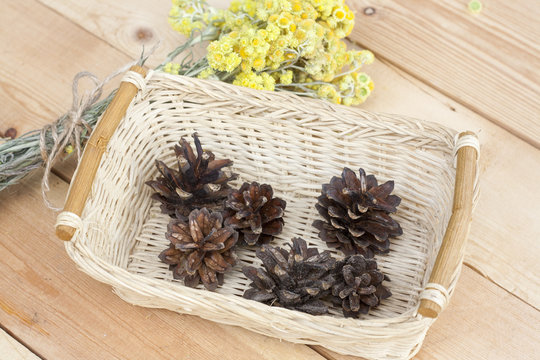 Dwarf everlast flowers bouquet and pine cones in a wicker basket on light wooden table, selective focus