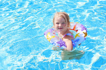 Fototapeta na wymiar Adorable happy little child, curly toddler girl in swimming suit having fun relaxing and floating on an inflatable toy ring in a pool on sunny day during summer vacation in resort