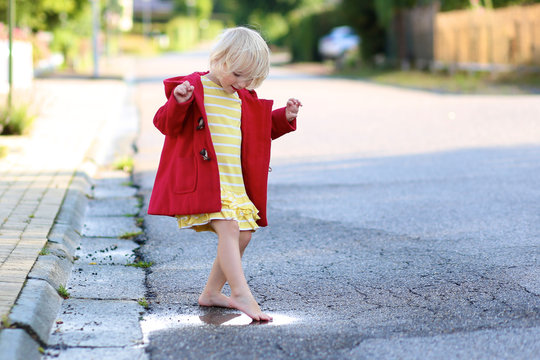 Happy Little Child, Adorable Blonde Curly Toddler Girl Wearing Red Duffle Coat Enjoying Sun After Rain Running Barefoot And Jumping On The Puddle On The Street On A Sunny Autumn Or Spring Day