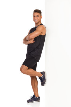 Man Rests Against The Wall. Smiling Young Man In Sport Black Shirt And Shorts Standing And Leaning On A Imagine Wall. Full Length Studio Shot Isolated On White.