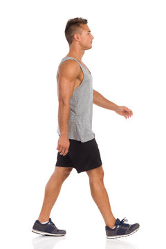 Young Man In Beige Shorts, Blue Shirt And Sneakers Walking. Side View. Full Length Studio Shot Isolated On White.