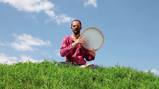 Young man sitting on the green grass and playing the tambourine in front of background of blue sky.
