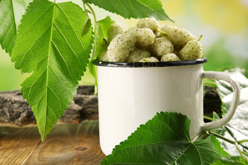 Ripe mulberries in mug with green leaves on table close up
