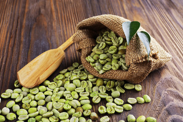 Green coffee beans with leaves and spoon in bag on wooden table close up