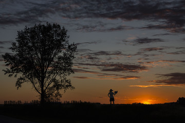 beautiful horizontal image of a silhouette of a lady wearing a dress and big hat standing close to a big tree reading a book under a gorgeous orange sunset in summer time