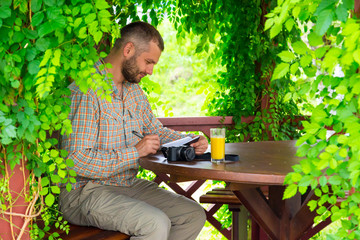 Travel diary.
Bearded man sitting at vintage design wooden arbor and writing notes into his notepad having glass of orange juice and photo camera on table surrounded green flora sun light background - Powered by Adobe