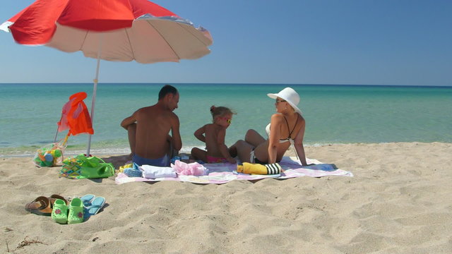Family on sand beach in shade of umbrella against turquoise water and clear sky
