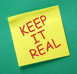 The phrase Keep It Real in red text on a yellow sticky note posted on a green notice board as a...