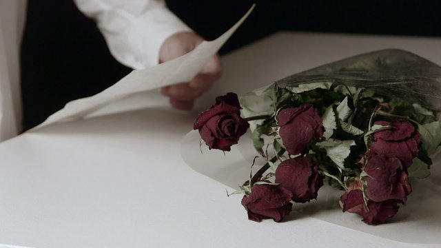 Man reading farewell letter with bouquet of withered roses on table
