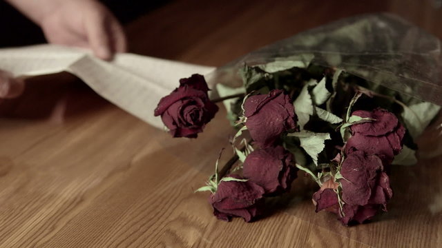 Man reading handwritten goodbye letter on table with wilted bouquet of roses
