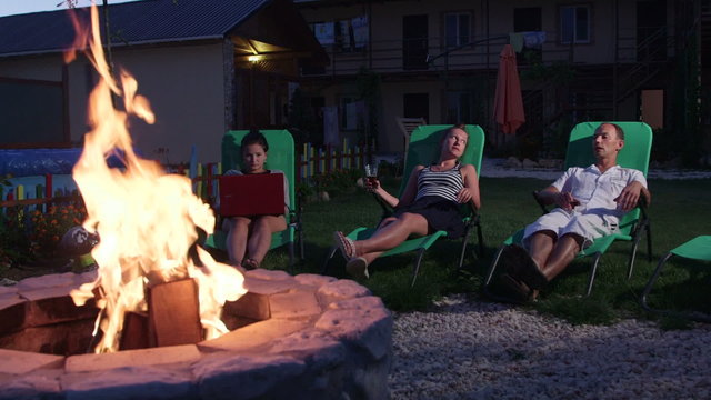 Friends having outdoor garden party with drinks relaxing near fire pit
