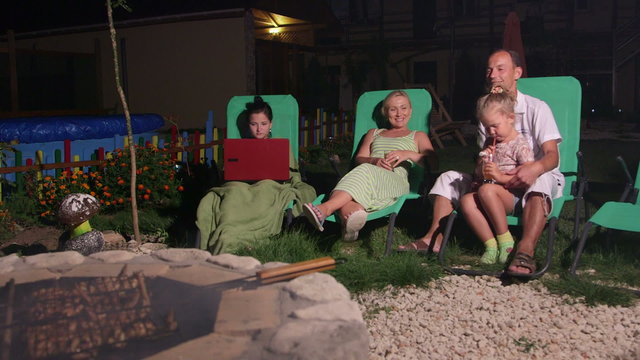 Family sitting on patio loungers grilling barbecue on stone fire pit in backyard
