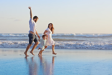 Positive family - father, mother with baby son hold hands and run with fun along edge of sea on smooth sand beach. Active parents and people outdoor activity on tropical summer holidays with children