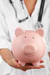 Doctor in white uniform with stethoscope holding pink piggy bank, closeup