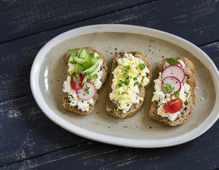 sandwiches with cream cheese, cucumber, egg, radishes and tomatoes on an oval dish on dark wooden background