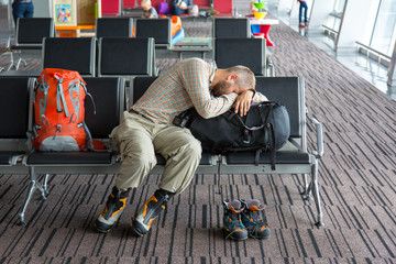 Airport lounge and people waiting for boarding.
Body of male on foreground sleeping on his luggage lying in chair other people miscellaneous actions on background terminal interior with large windows