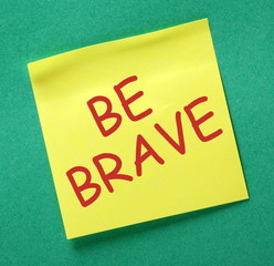 The phrase Be Brave in red text on a yellow sticky note posted on a green notice board as a reminder