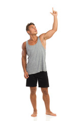 Smiling muscular man standing barefoot, looking up, holding arm raised and pointing up. Full length studio shot isolated on white.