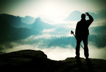 Fototapeta premium Misty day in rocky mountains. Silhouette of tourist with poles in hand. Hiker stand on rocky view point above misty valley.