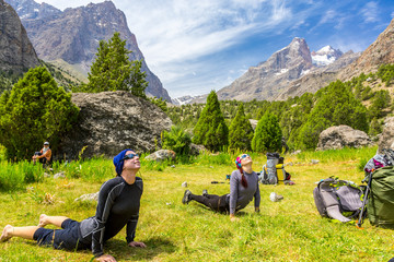 Young women doing morning fitness in mountain landscape.
Two female athletes on green grass lawn doing yoga sport exercises outdoor day sunlight blue sky and mountains on background