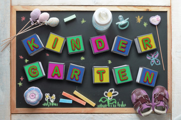 Some letter cubes forming the word kindergarten, a baby bottle, a pacifier, some toys and sneakers on a black chalkboard from above. Children education related objects.