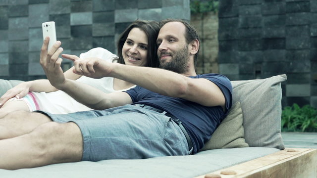 Happy couple taking selfie photo with cellphone lying on daybed
