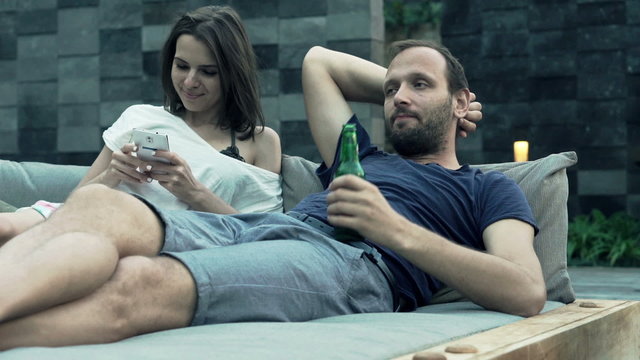 Young couple lying on daybed, woman using smartphone and man drinking beer 
