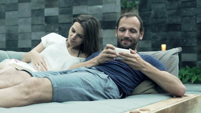 Young couple lying on daybed, woman reading book and man playing on smartphone 
