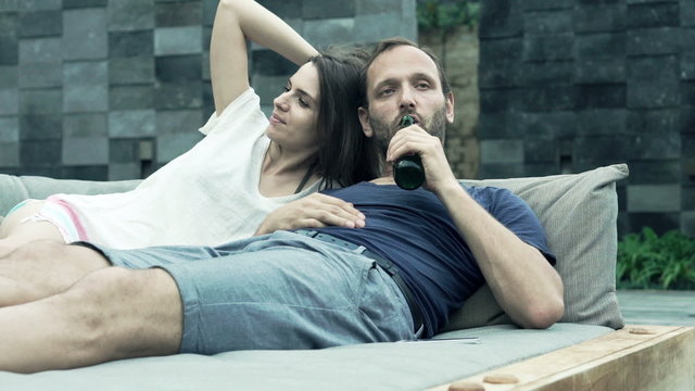 Young couple talking and drinking beer while lying on daybed
