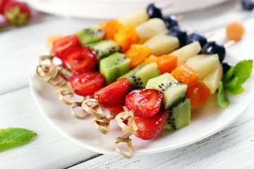 Fresh fruits on skewers in plate on wooden table, closeup