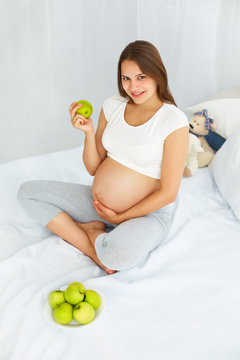 Pregnant Young Woman holding Apple while sitting on the Bed. Hea