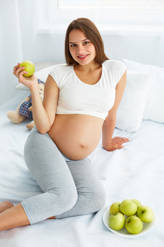 Pregnant Young Woman holding Apple while sitting on the Bed. 