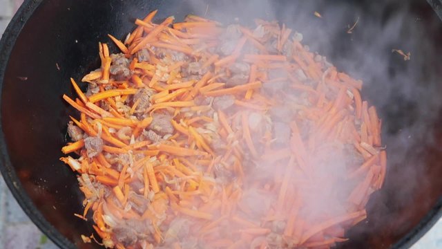 Uzbek national dish pilaf in a large cast-iron cauldron on the fire, add sliced red carrots, rice, lamb, mutton fat, the fat tail, sesame oil, onion, garlic, red hot chili pepper