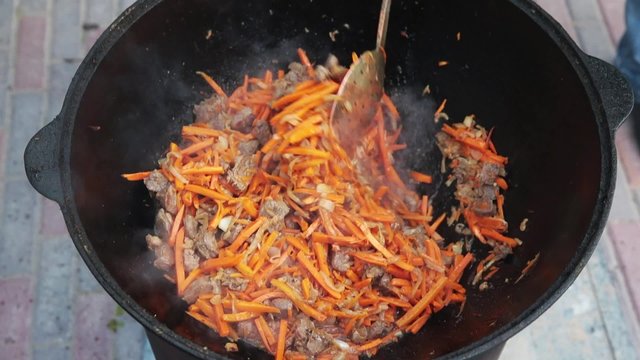 Uzbek national dish pilaf in a large cast-iron cauldron on the fire, add sliced red carrots, rice, lamb, mutton fat, the fat tail, sesame oil, onion, garlic, red hot chili pepper
