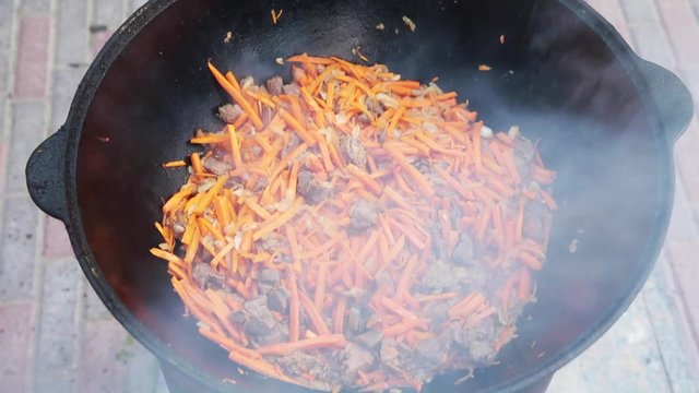 Uzbek national dish pilaf in a large cast-iron cauldron on the fire, add sliced red carrots, rice, lamb, mutton fat, the fat tail, sesame oil, onion, garlic, red hot chili pepper