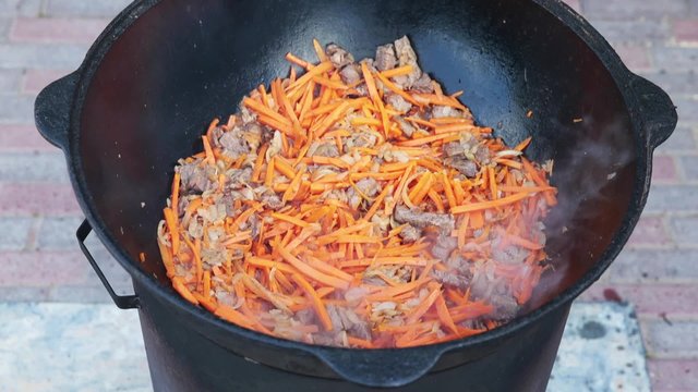 Uzbek national dish pilaf in a large cast-iron cauldron on the fire, add sliced red carrots, rice, lamb, mutton fat, the fat tail, sesame oil, onion, garlic, red hot chili pepper