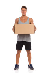 I Have Delivery For You. Smiling young man in black shorts and sneakers standing with legs apart and holding carton box. Full length studio shot isolated on white.