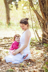  pregnant mother in white dress sitting on ground in park outside with daughter in pink clothes on her laps  on sunny spring summer day 
