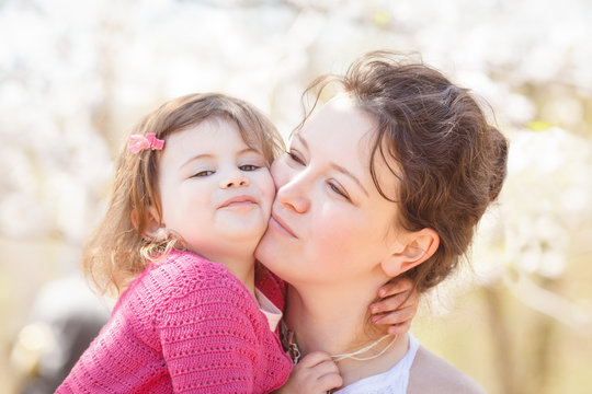 Close up portrait of caucasian mother in white dress holding hugging her daughter in pink clothes  kissing her in cheek on sunny spring summer day in the park outside 