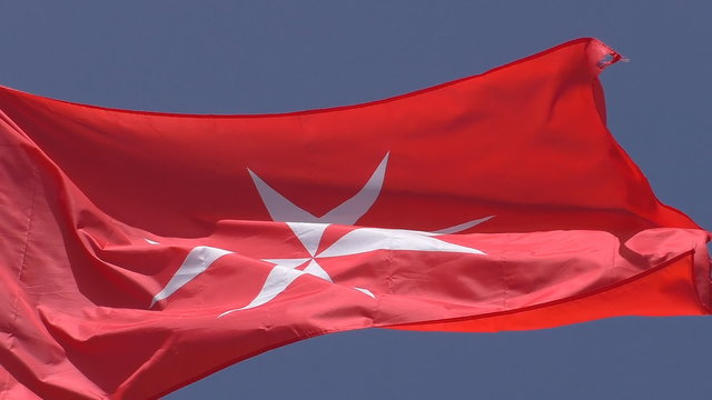 Close up of white Maltese cross on red background flag, the national flag of Malta or civil ensign flown by civil ships, isolated against the blue sky waving in a breeze     