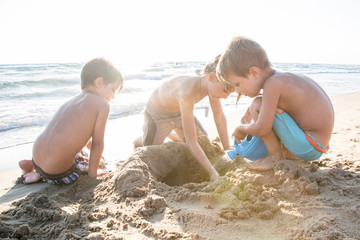 Tre bambini che giocano con la sabbia in riva al mare, controluce - obrazy, fototapety, plakaty
